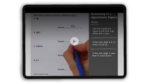 A hand holding a pen points at a digital tablet displaying a math rounding lesson, with text and example numbers visible on screen.