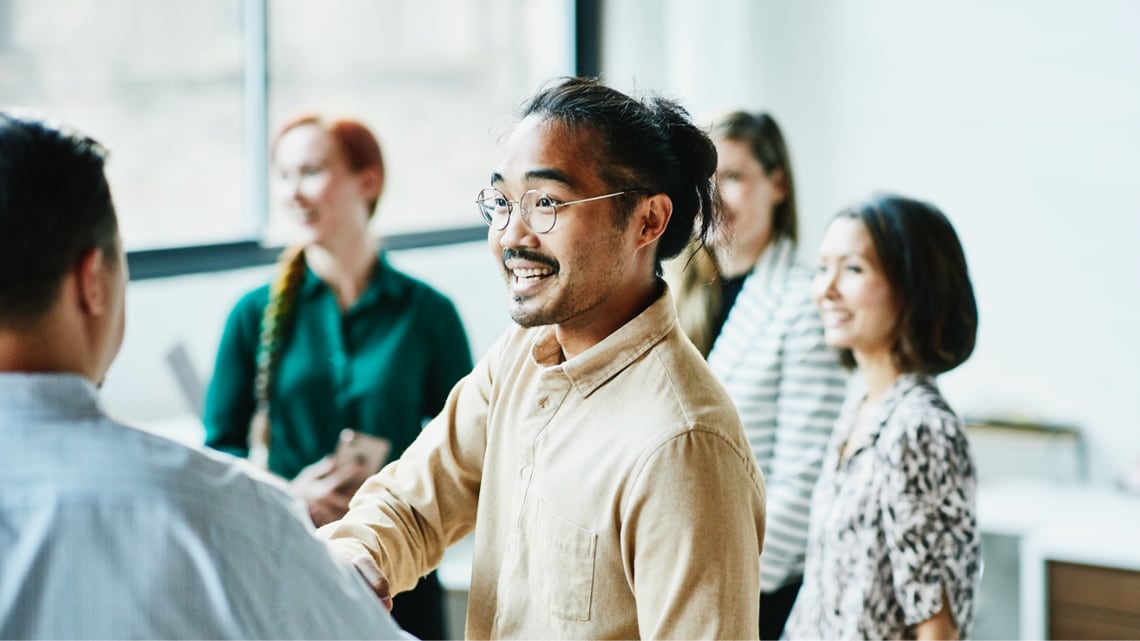 Group of colleagues greeting each other in a bright office space, with one person shaking hands while others stand nearby engaged in conversation.