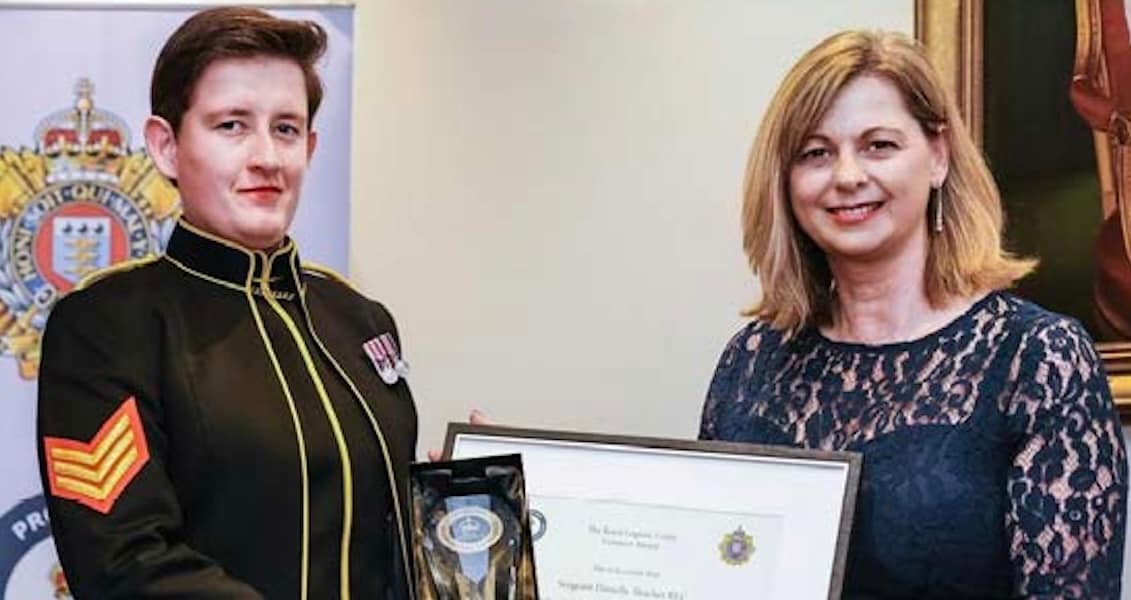 Award presentation scene with a uniformed individual receiving a framed certificate and trophy from another person at an official ceremony.
