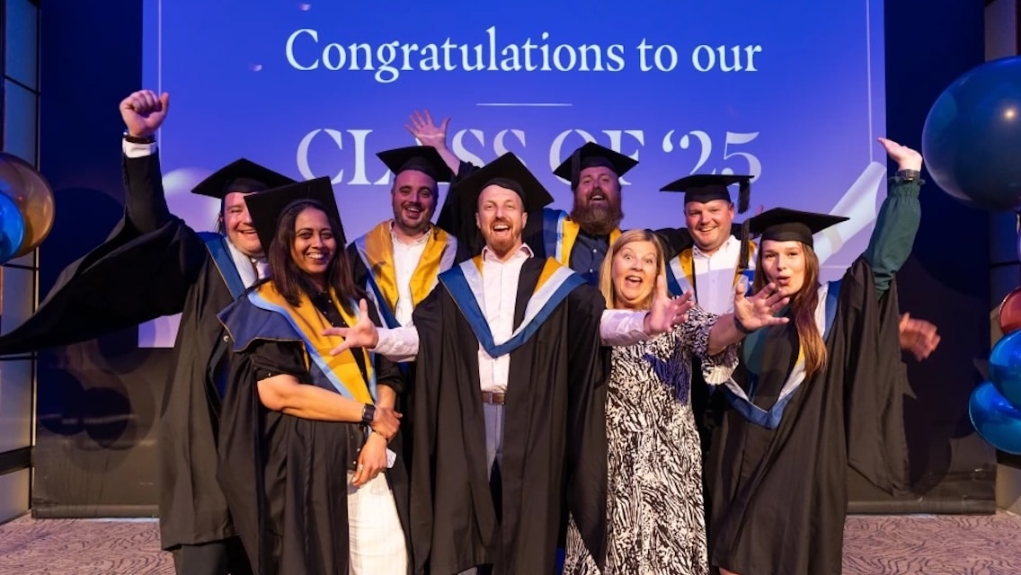 Group of graduates in caps and gowns celebrating together on stage in front of a banner that reads “Congratulations to our Class of ’25”.