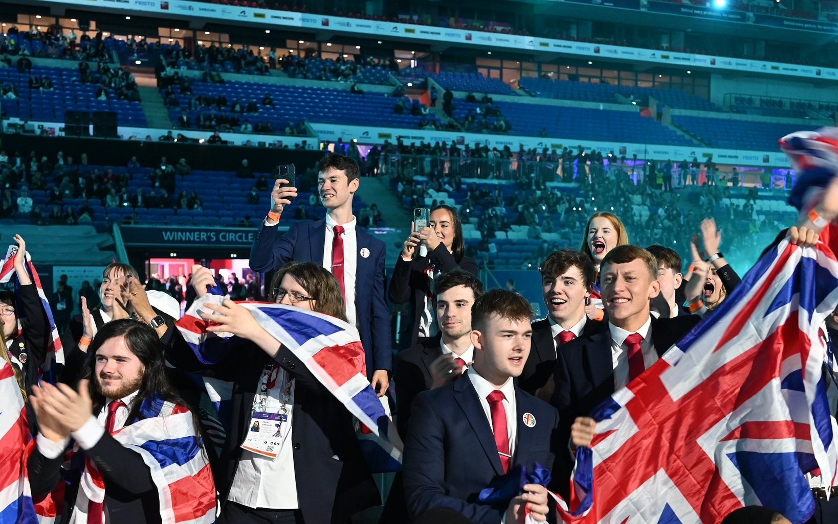 Team UK, many dressed in formal attire with red ties, enthusiastically wave large Union Jack flags in a crowded arena