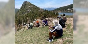 A group of six hikers sitting on a hillside in a hilly area with a lake and mountain peak in the distance.