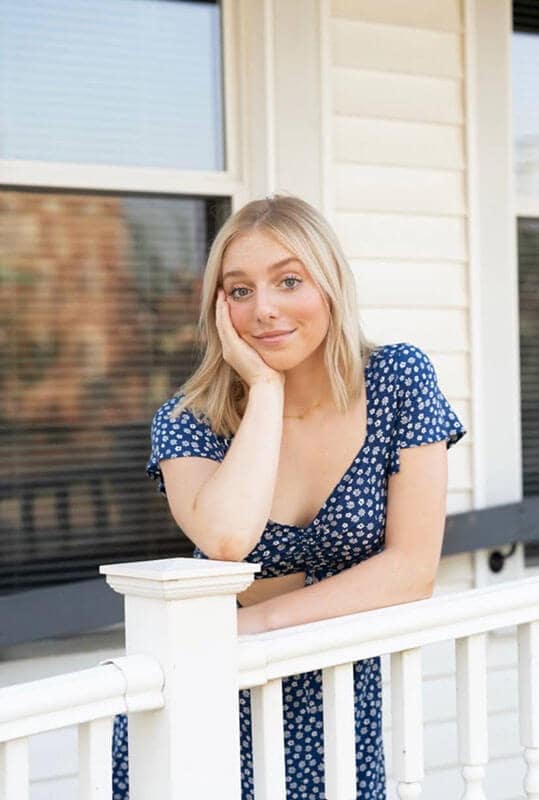 Blog author Allie is leaning against a white railing resting her chin in her hand. She has straight blonde hair and is wearing a blue and white printed dress.