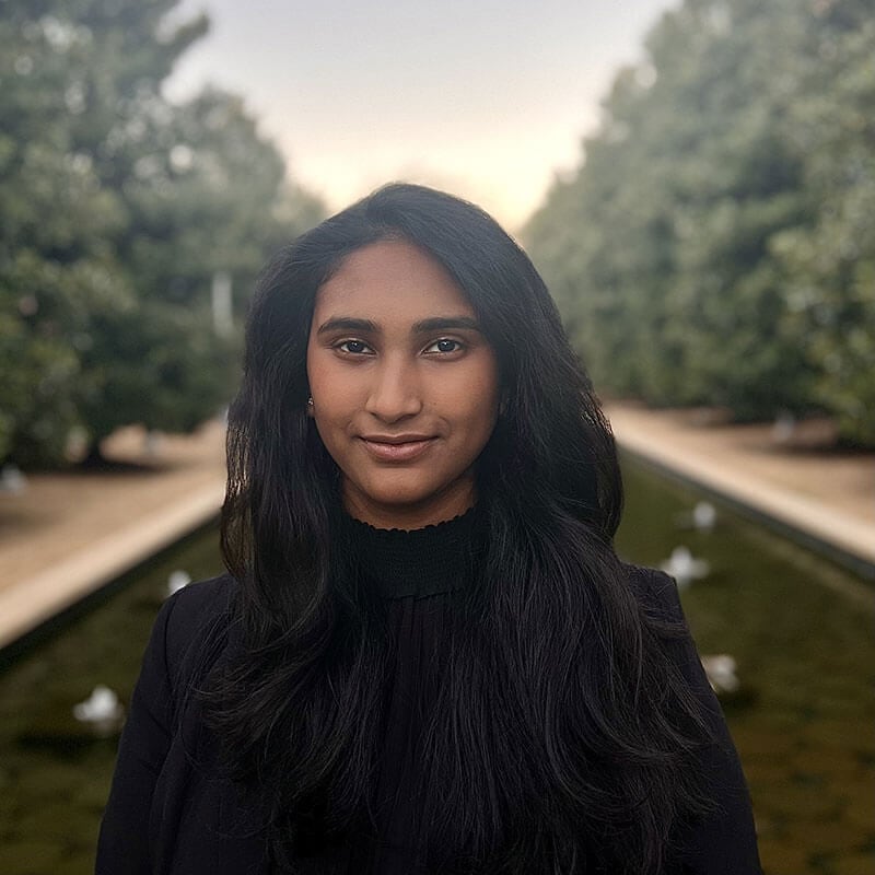 Blog author Akshaya has long dark straight hair and is wearing a dark blazer over a dark turtleneck. Akshaya is standing outside in front of two rows of trees.