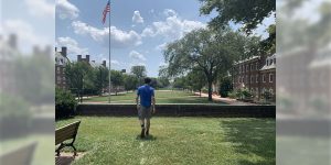 A young male college student walking across a college campus quad. He is wearing a blue t-shirt and grey shorts.