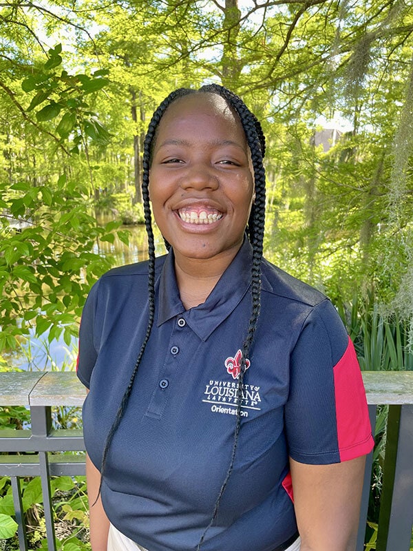 Blog author Ariyannah is standing outside in front of a wooden railing. She has long dark braids and is wearing a blue polo shirt with her college logo on the pocket area.