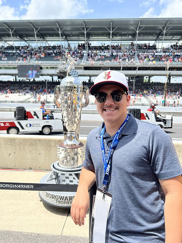 Blog author Aj is at a racetrack standing in front of the trophy awarded for the Indianapolis 500.