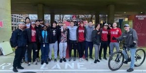 A group of 20 college men gathered outside wearing hoodies, jackets, and hats. One young man is on a bike.