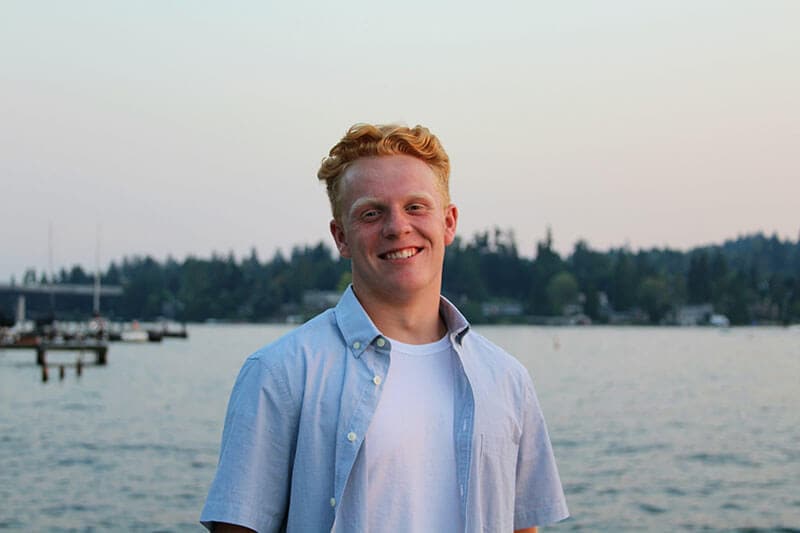 Blog author Bryson is standing outside in front of a lake. He has short red hair and is wearing a short-sleeved button shirt open over a white t-shirt.