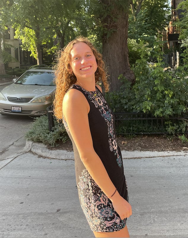 Blog author Cara is standing outside on a campus sidewalk. She has long red, curly hair and is wearing a black sleeveless dress.