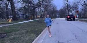 A young woman jogging down a neighborhood street. She is wearing a long-sleeved blue t-shirt, black shorts, and light orange running shoes.