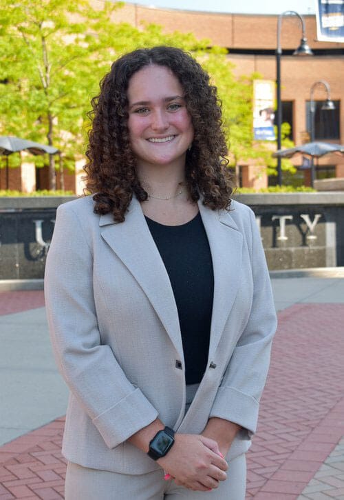 Blog author Carla has shoulder length curly hair. She is standing outside on campus wearing a light-colored blazer over a dark blouse.