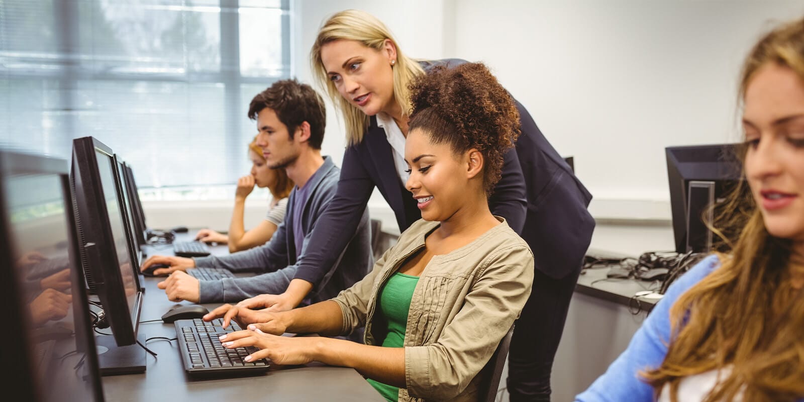 Group of individuals sitting in a computer lab while an instructor discusses information on device.