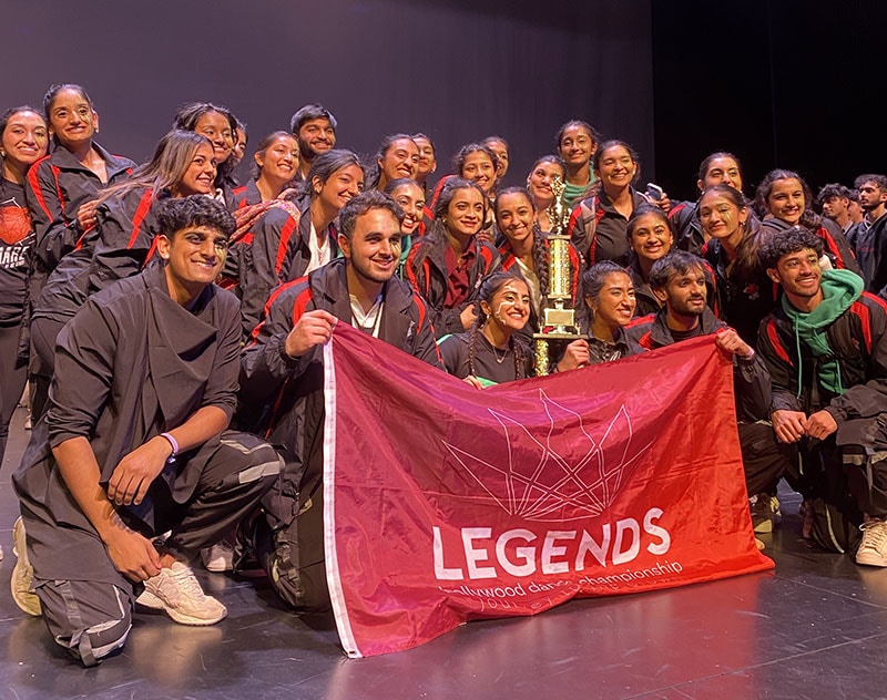 The blog author’s Bollywood dance team in a group photo on stage at the Legends competition. They are holding a red flag and a trophy.