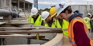 Four construction management students at Texas A&M wearing hard hats and looking down into a concrete structure.