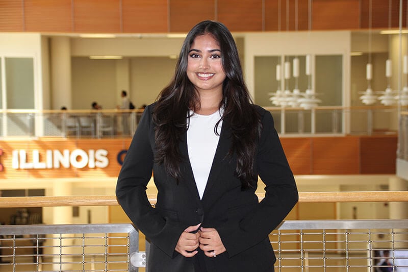 Blog author Keya is standing by a railing in a building on her campus. She has long, straight, black hair and is wearing a black blazer over a white top.