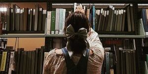 A female college student with her hair pulled back in a bow barrette is reaching for a book from a library shelf.