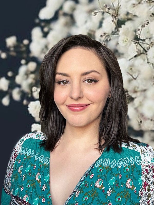 Blog author Lindsay has medium length dark hair. She is standing in front of a white flowering tree and is wearing an emerald green print blouse.