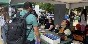 A college student wearing a backpack speaks with two staff members at a college organization fair.