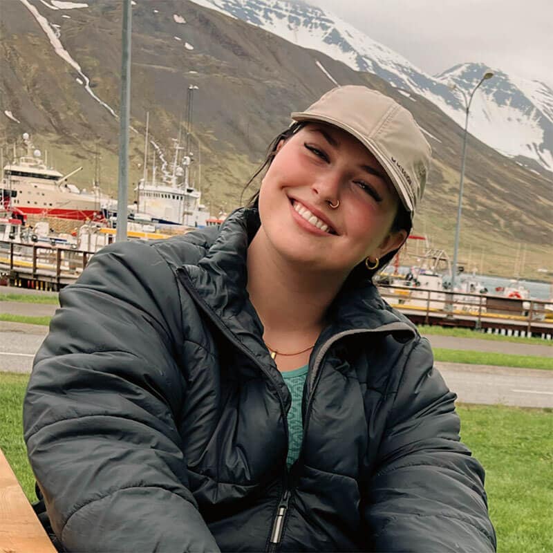 Blog author Maggie is sitting outside with snow-capped mountains in the distance. She is wearing a black puffy coat and a tan baseball hat.