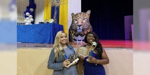 Two collegiate women each holding a trophy, stand in front of a stage with a large tiger cut-out and blue and yellow decorations.