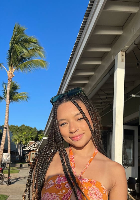 Blog author Olivia is standing outside on a sunny day with a palm tree in the background. She is wearing an orange halter top, has sunglasses on her head, and has long dark braids.
