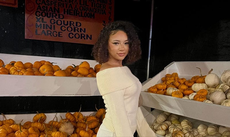 Blog author Olivia is standing in front of displays of mini-pumpkins. She has medium length curly dark hair and is wearing a cream-colored sweater.