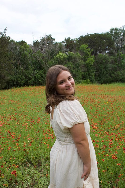 Blog author Peyton is standing outside in a meadow. She has medium length light brown hair and is wearing a white dress.