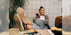 A college woman is smiling and sitting with her feet on a desk with her mobile phone in her hand.