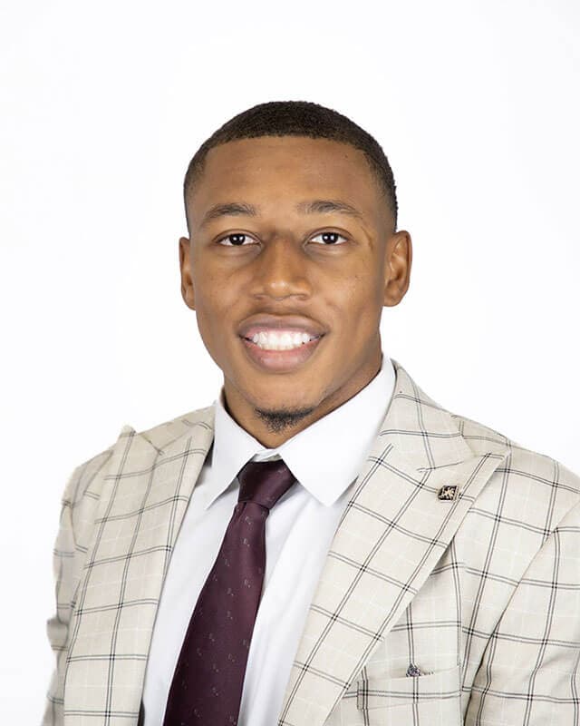 A studio headshot of blog author Royale. He has short dark hair and is wearing a light checkered blazer over a white shirt and maroon tie.