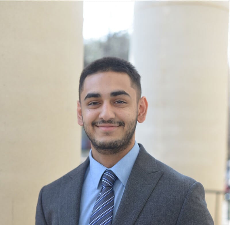 Blog author Sumay has short dark hair and is wearing a grey business suit, light blue shirt, and striped tie. He is standing outside in front of two large pillars.