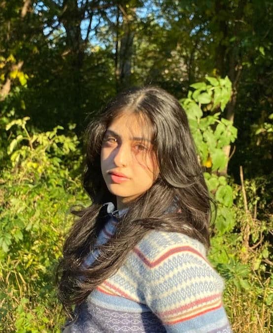 Blog author Shrimai is outside in front of green shrubbery. She has long dark hair and is wearing a multi-colored striped sweater.