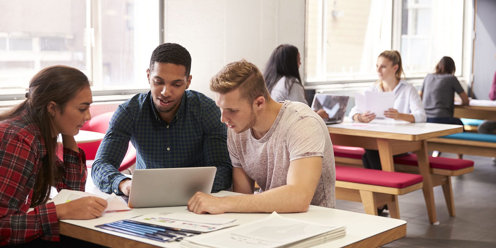 Three students sit at a table in a college common room. They are gathered around a laptop computer and are looking at the screen with interest.