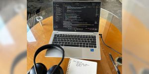 A laptop with coding language on the screen. A set of headphones and a spiral notebook sit nearby.