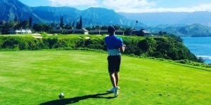 A young man swings a golf club on a course overlooking mountains and a body of water.