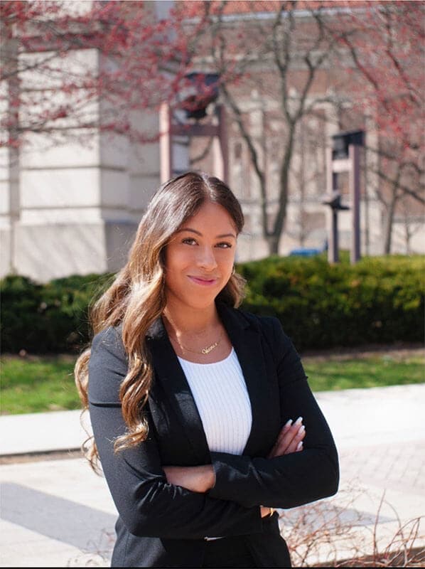 Blog author Victoria is standing outside on campus with her arms crossed. She has long dark wavy hair and is wearing a black blazer over a white blouse.