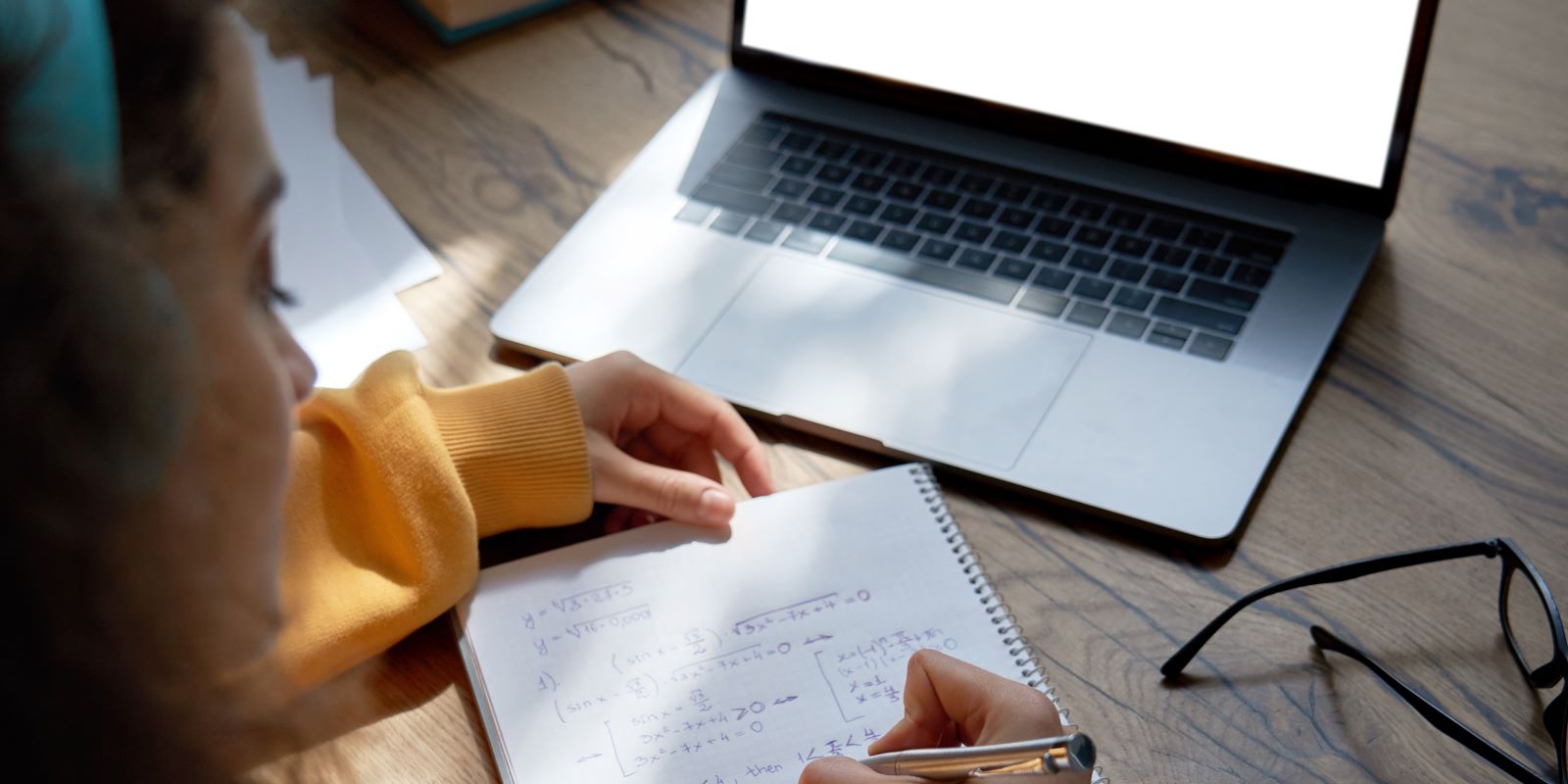 Image of student taking notes in a notebook, while reviewing content on their laptop