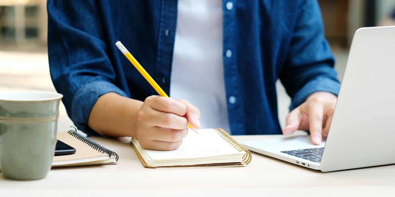 College-aged student with paper and pencil, writing in front of a laptop computer.