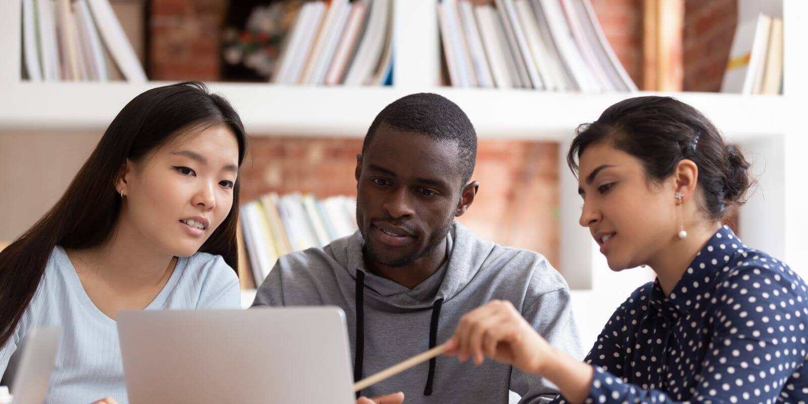 A diverse group of students sitting in front of shelves of books, focused on a laptop.