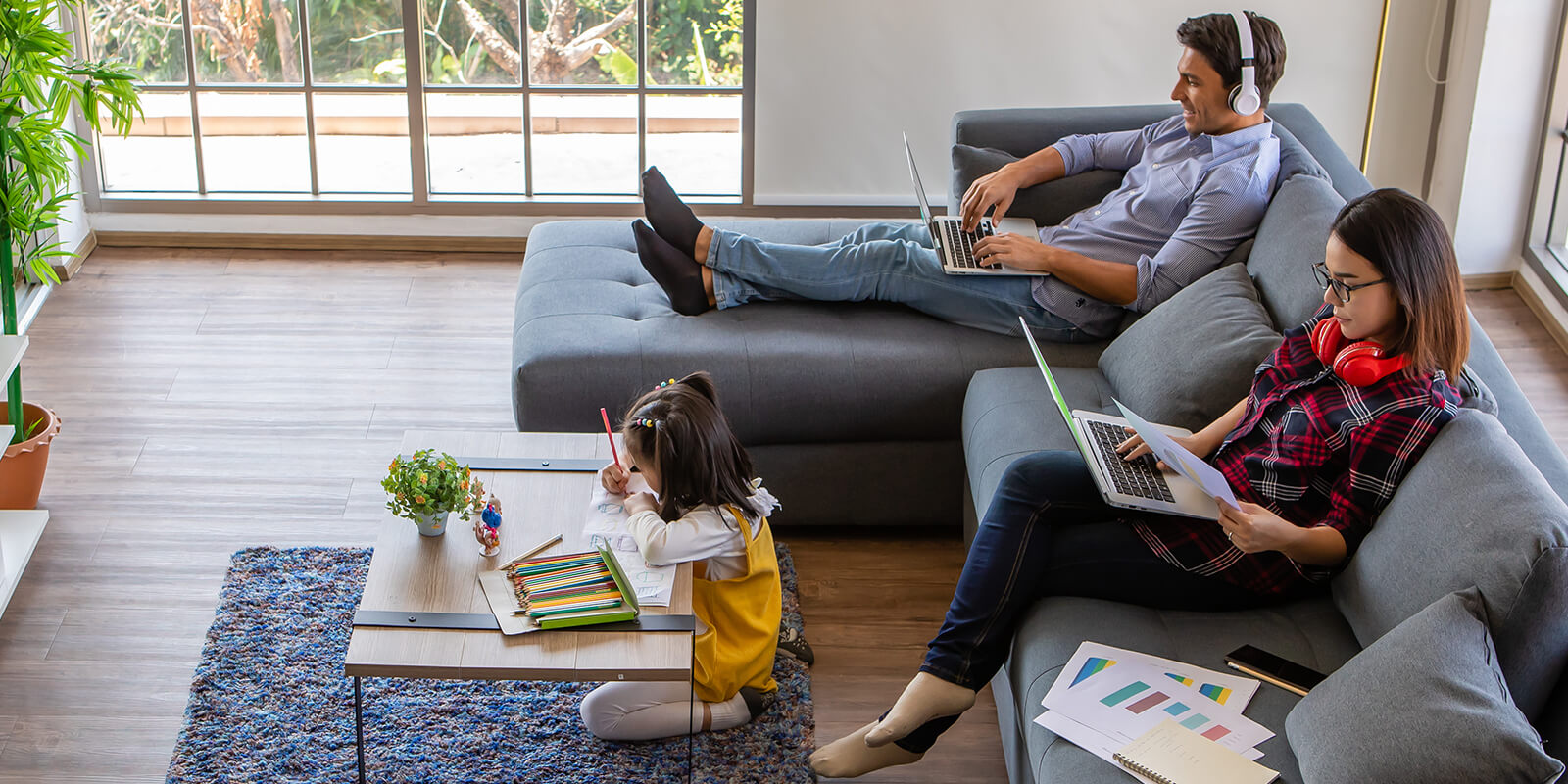 A man with headphones on and a woman in a plaid shirt sit on a gray couch typing on laptops while a little girl in a yellow dress kneels over a coffee table drawing with colored pencils.