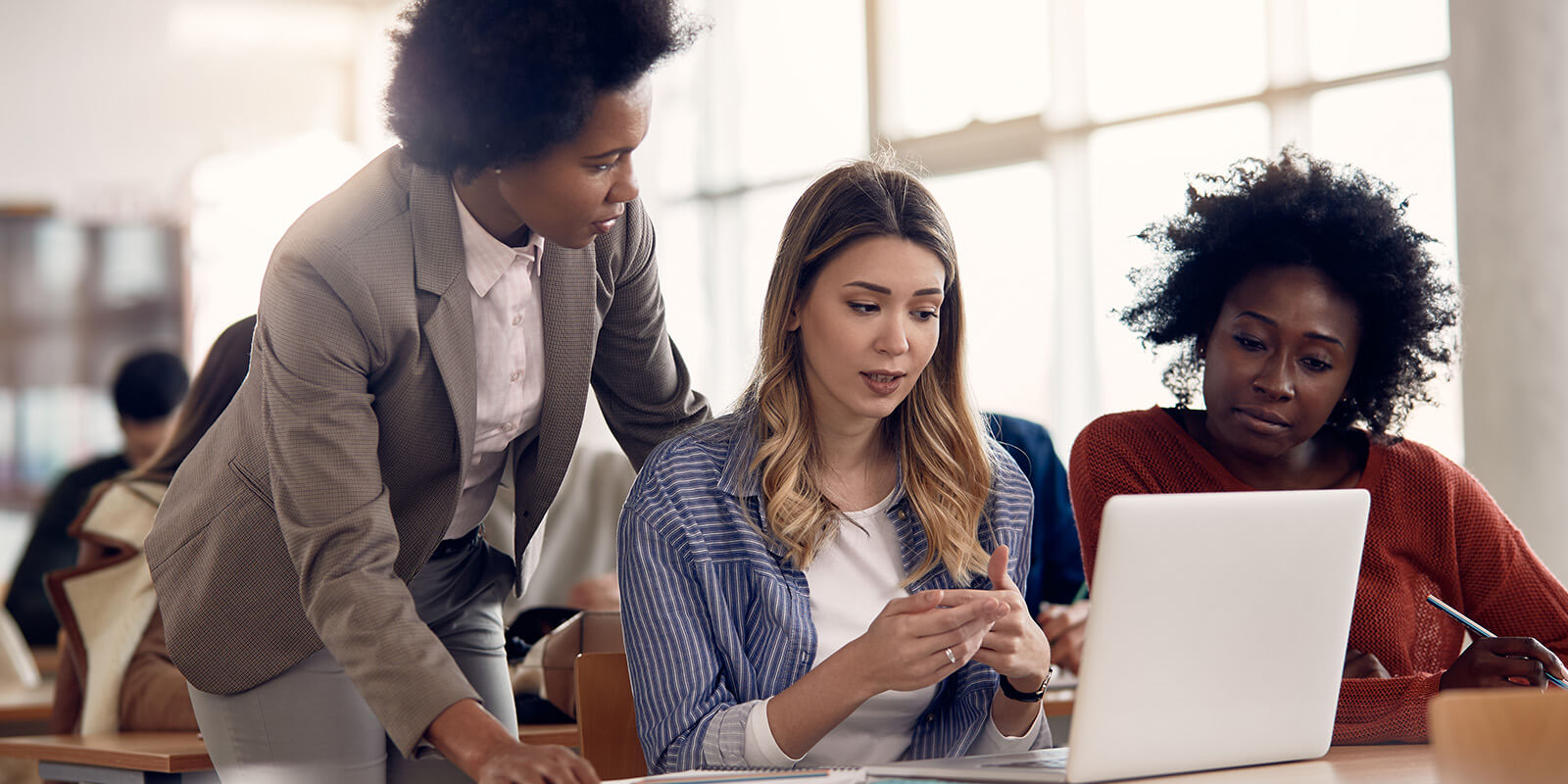 Three women are looking at a laptop computer screen. Two are seated at a desk while the third is standing.