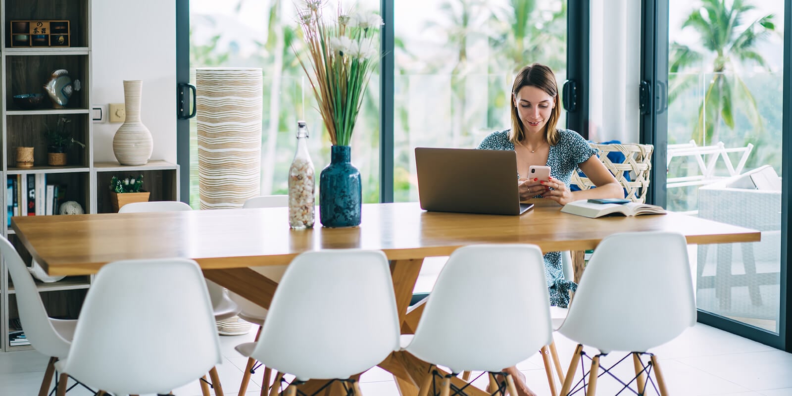A woman sitting at a table, looking at phone, laptop on the table.