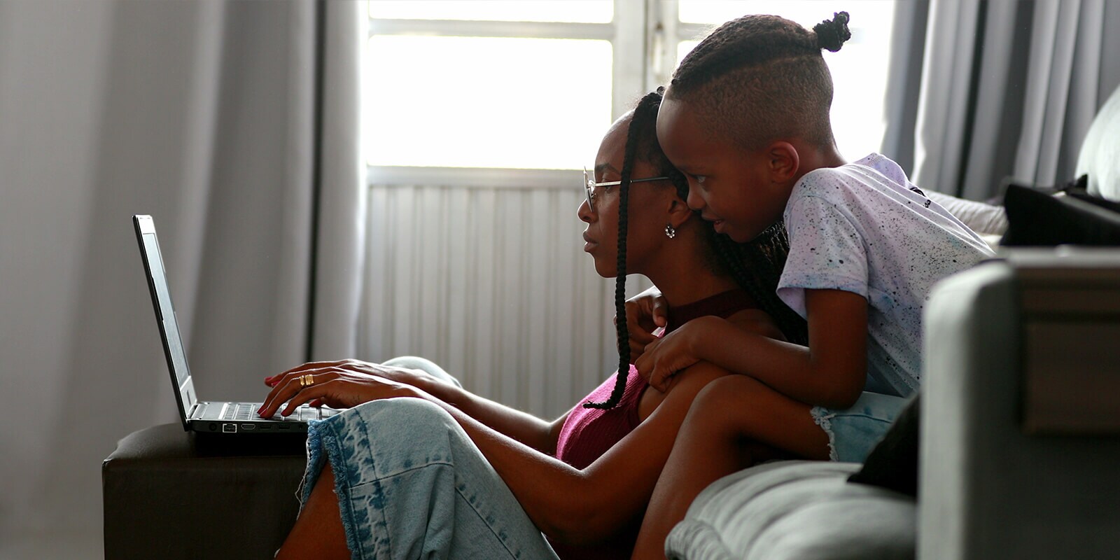A woman working on laptop sitting on the floor, a child sitting on a couch looking at the laptop screen behind her.