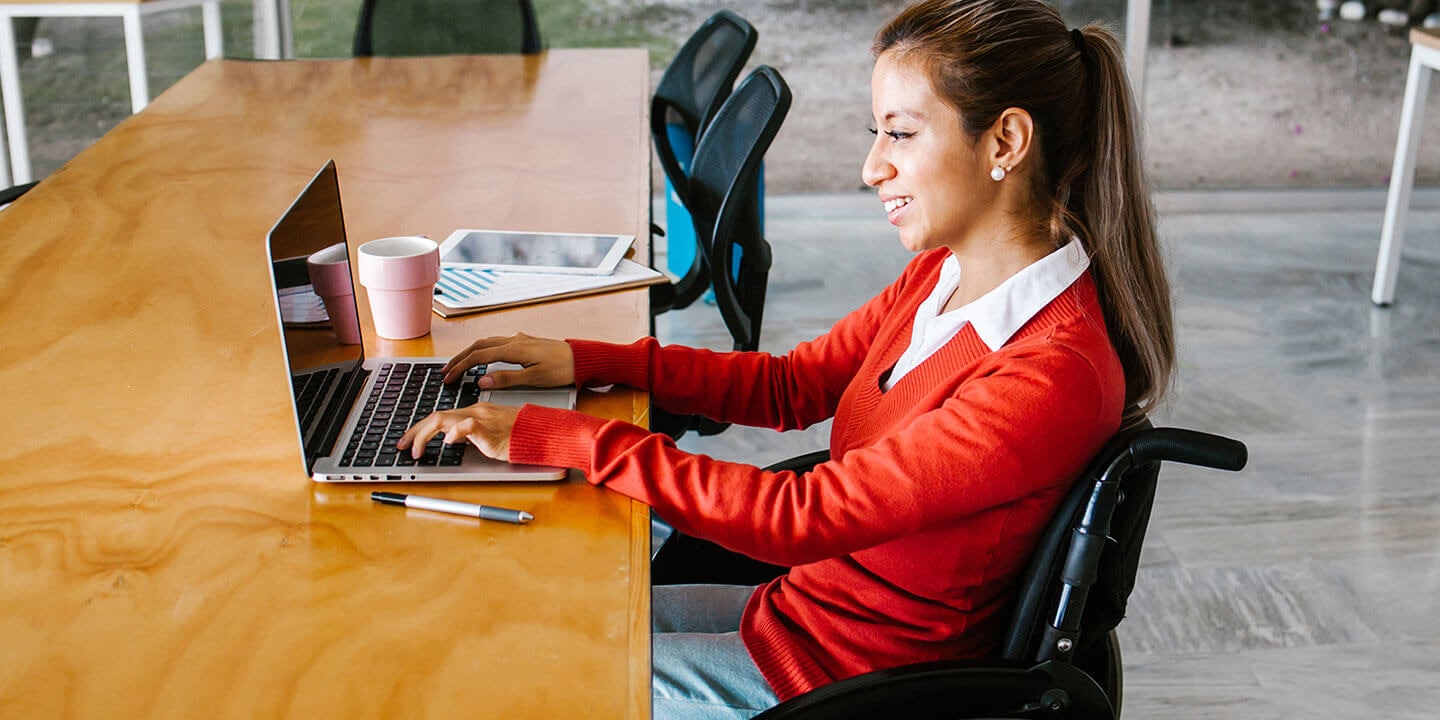 A woman is sitting at the table and working on a laptop. Apen, cup and stationary are on the table.