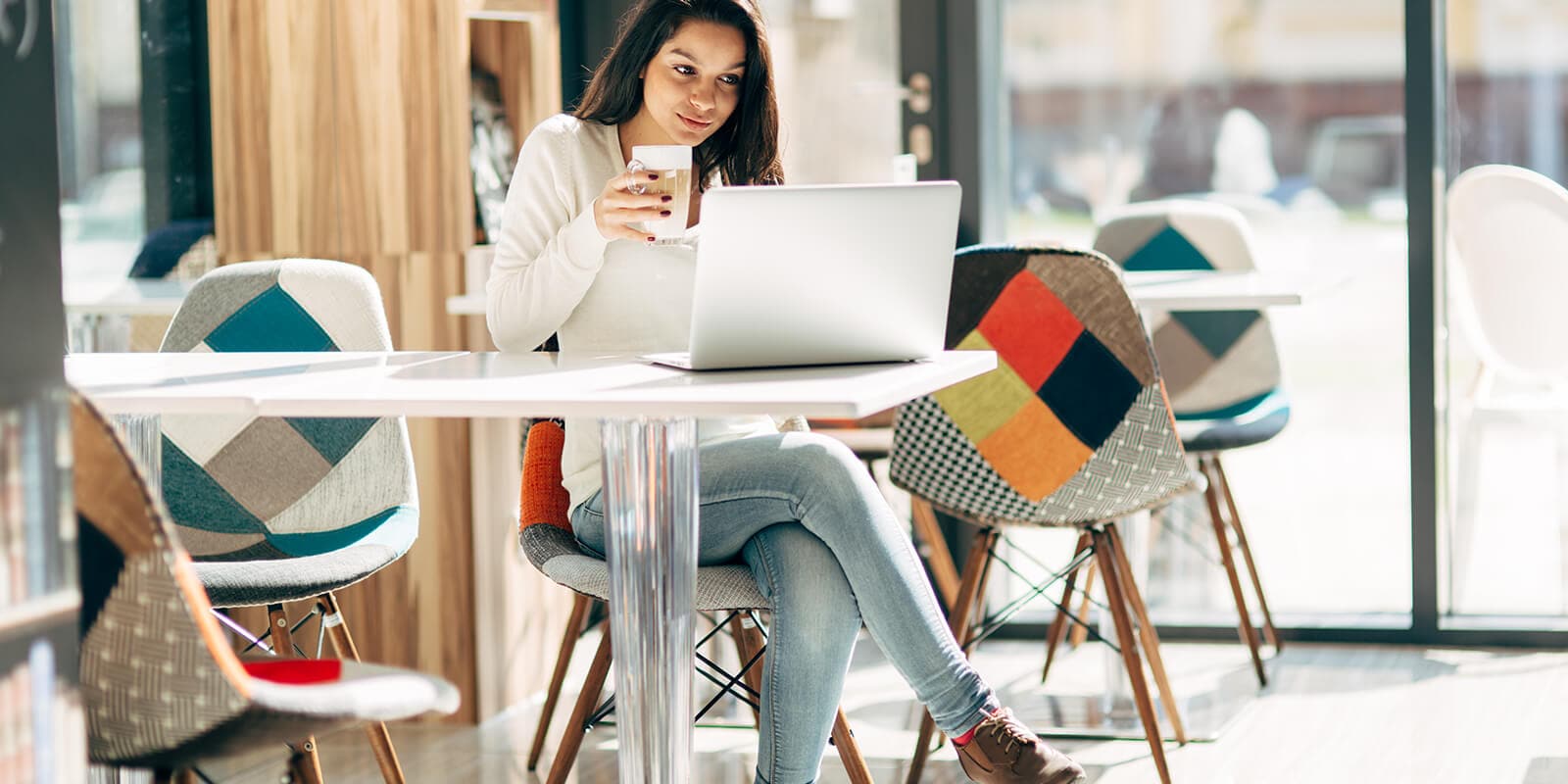 A woman looking sitting at a table, looking at the laptop with a coffee cup in her hand.
