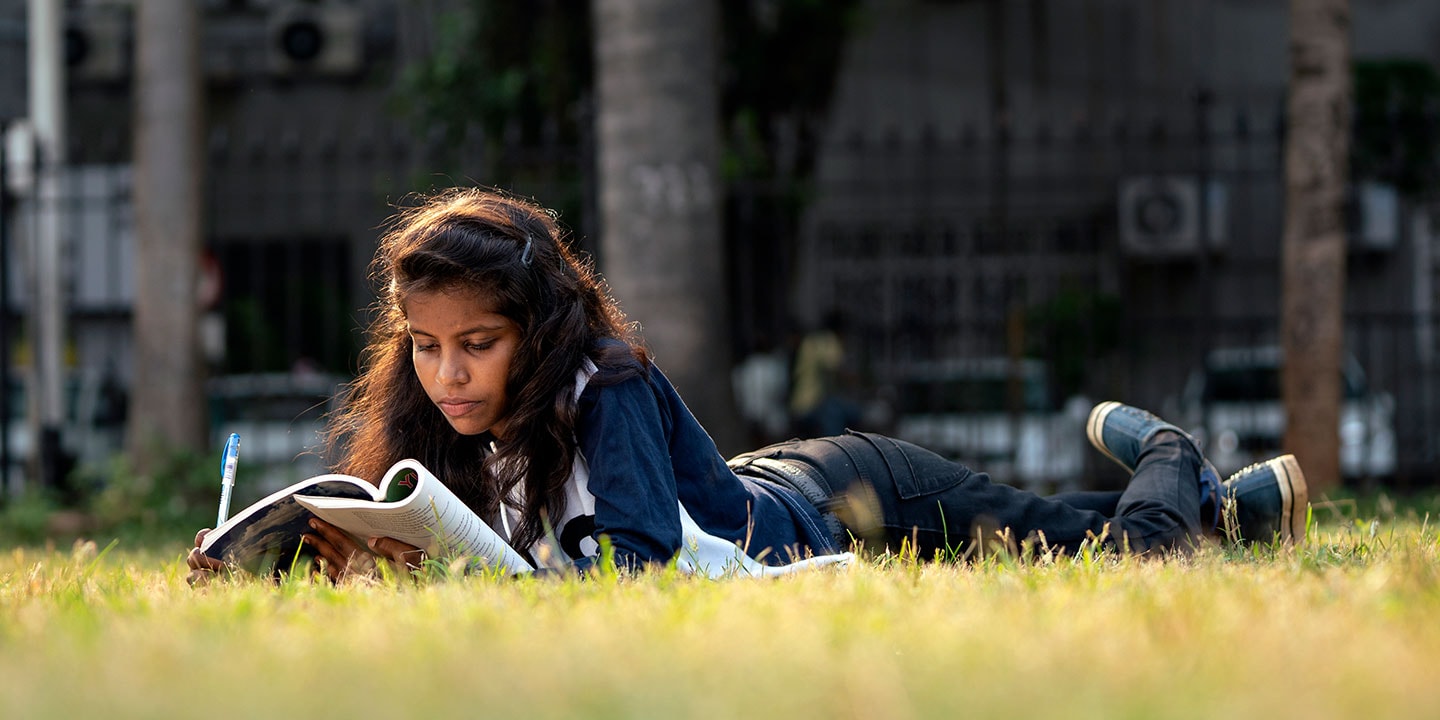 A female student lies outside in the grass, propped up on her elbows and reading a book.