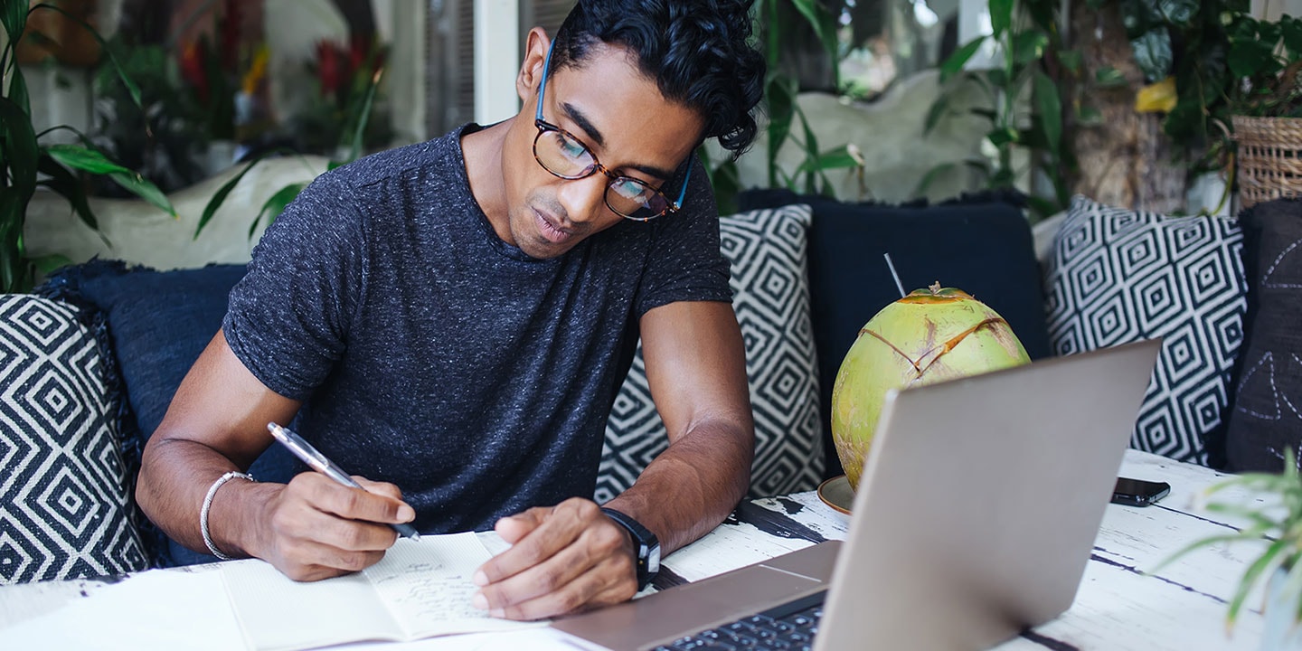 A man sits at a table, writing in a notebook, with a laptop computer open in front of him.