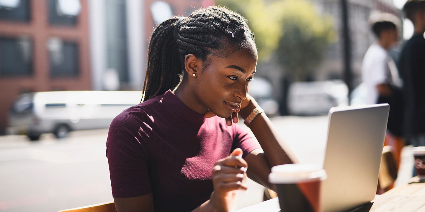 A woman sits outside at a cafe, looking at a computer. A cup of coffee is next to her.