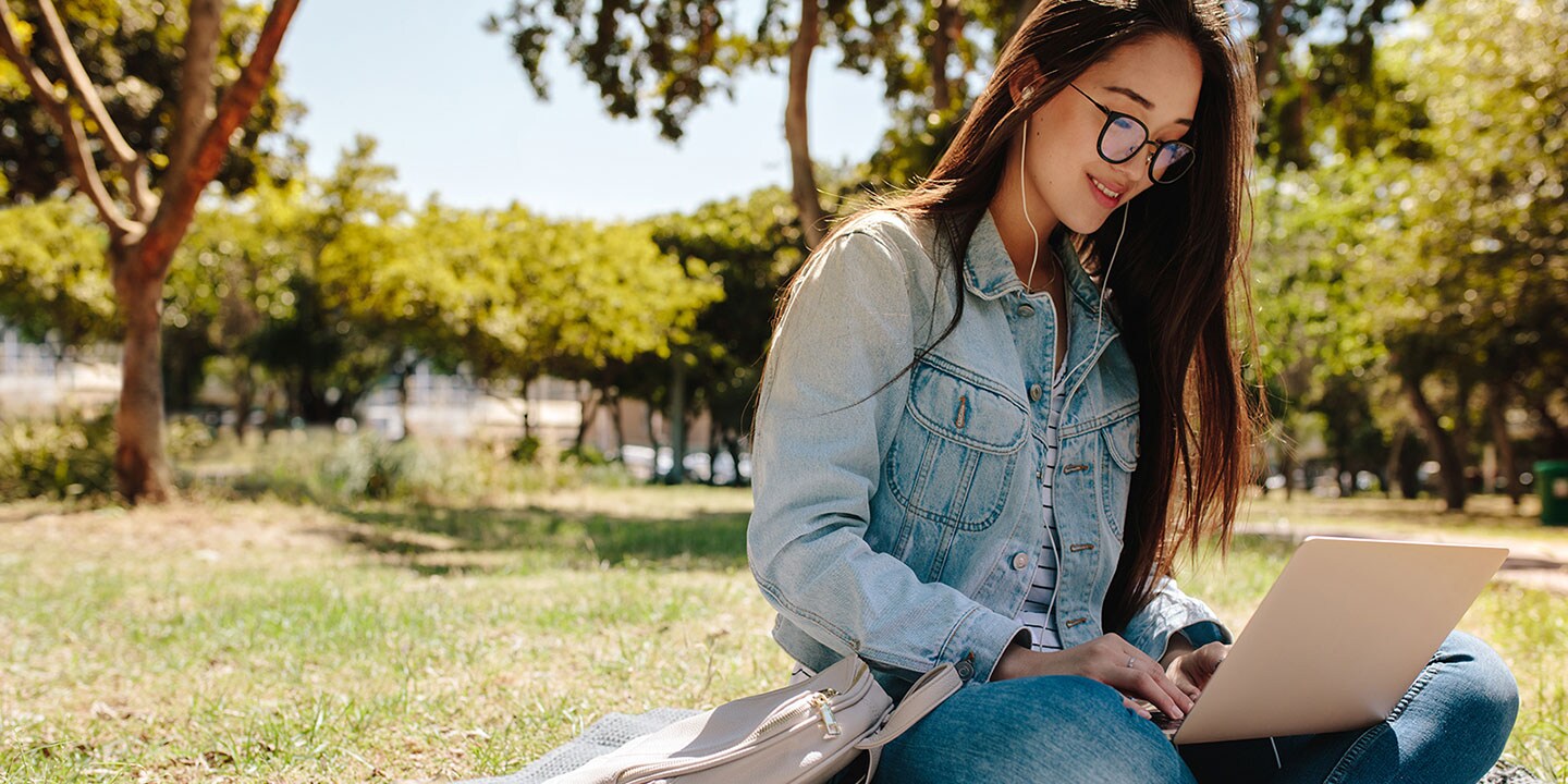 A woman is sitting on the ground outdoors, working on a laptop computer and wearing in-ear headphones.