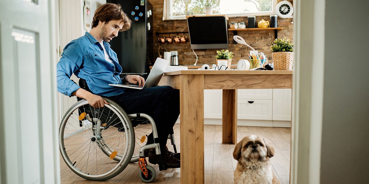 A man in a wheelchair sits at a kitchen table, working on a computer. A dog sits nearby under the table.